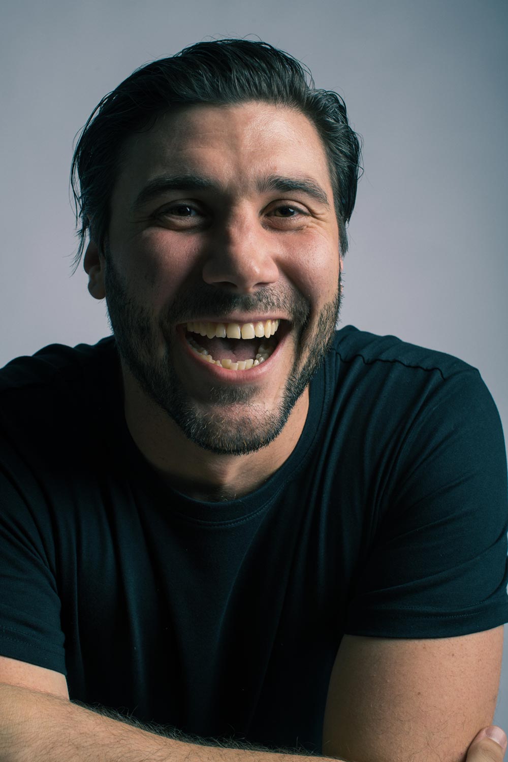 Headshot of a man wearing a black t-shirt, he is looking towards the camera laughing